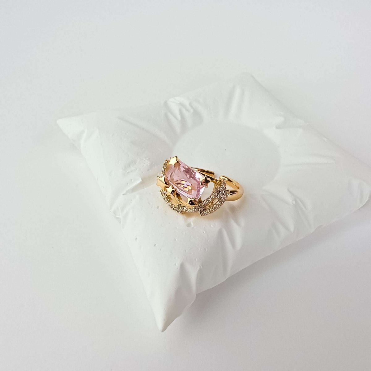 Позолоченное кольцо с розовым кристаллом и звёздными деталями на белой декоративной подушке